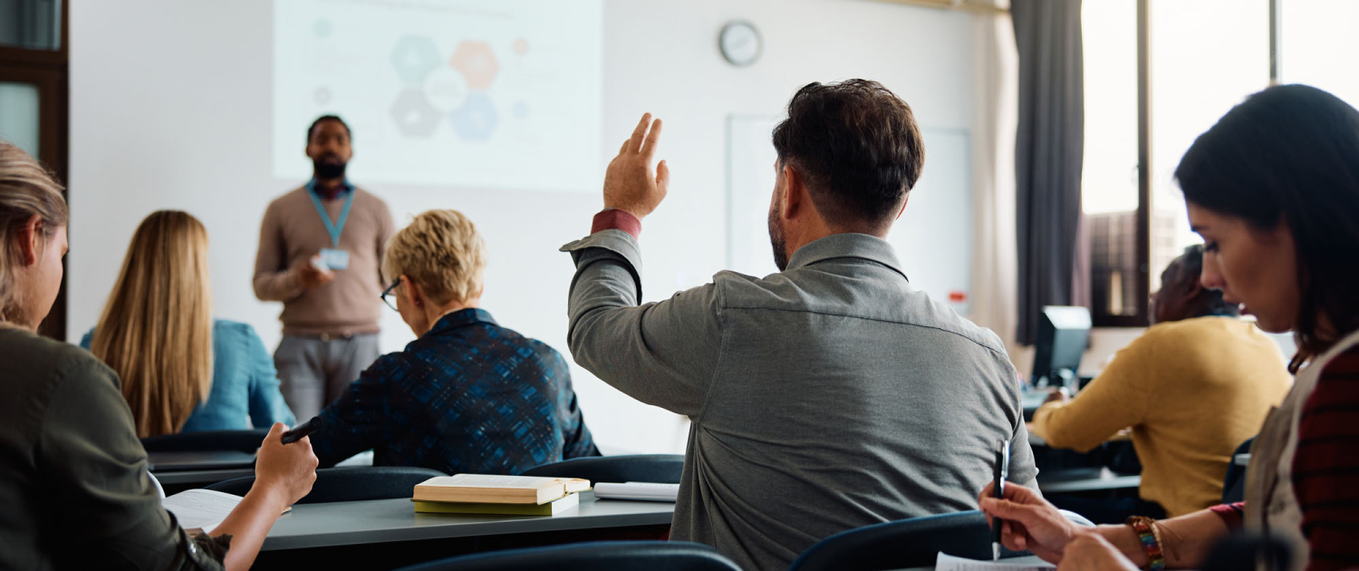 Back view of older student raising his hand to answer teacher's question during education training class.